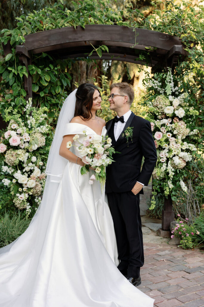 Bride and groom smiling under the floral-covered arbor during golden hour at their McCormick Home Ranch Wedding in Camarillo, CA.