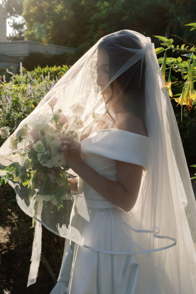 Bride standing in soft golden light with her veil draped over her bouquet at McCormick Home Ranch.