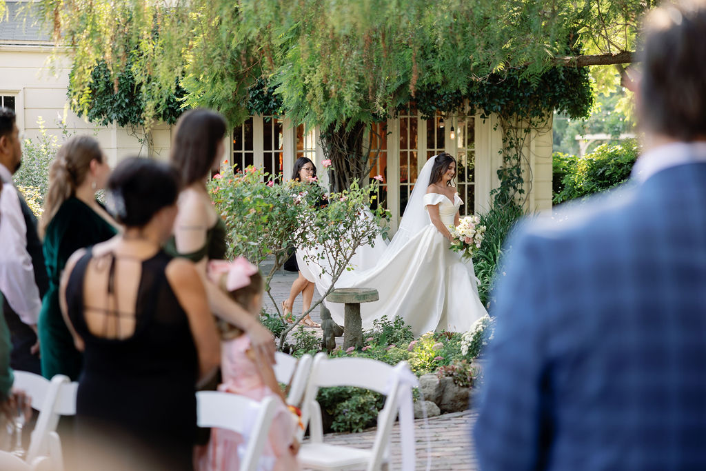 Bride walking toward the ceremony aisle with guests standing and watching in the garden setting.