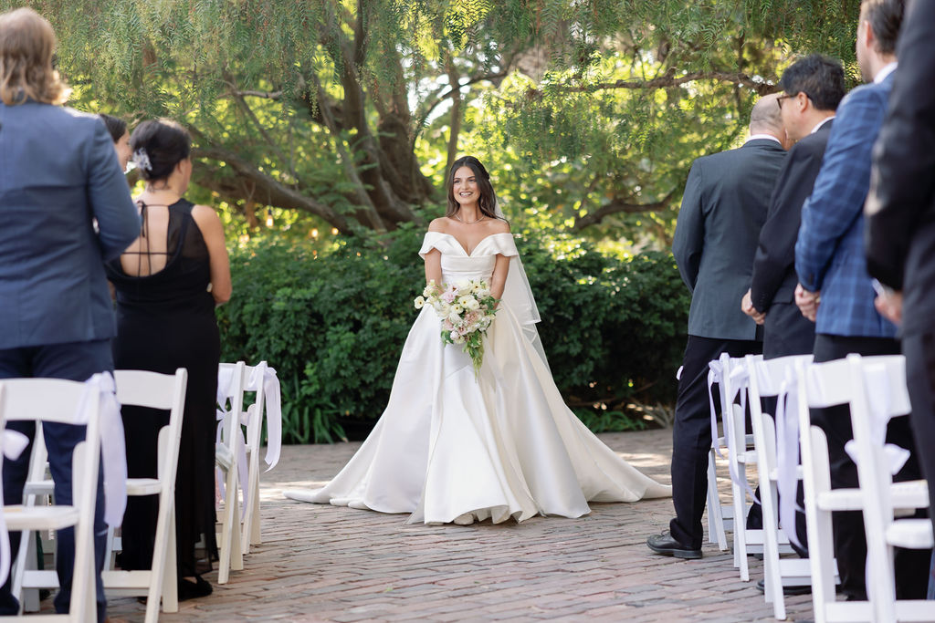 Bride walking toward the ceremony aisle with guests standing and watching in the garden setting.