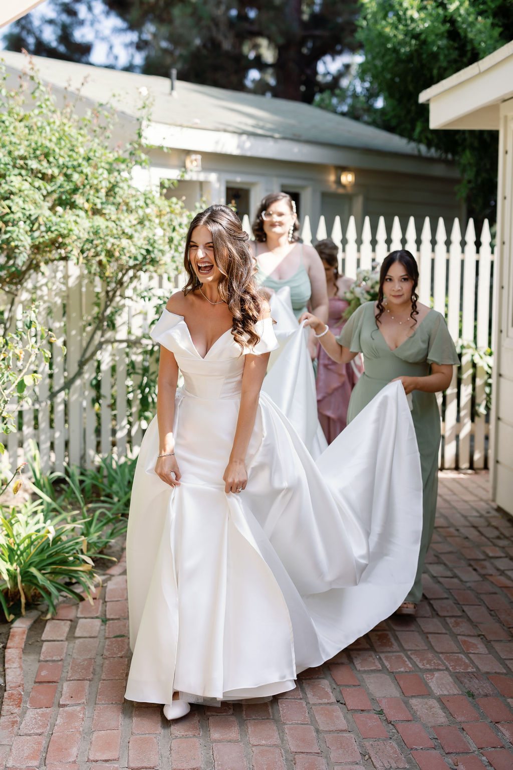 Bridesmaids helping arrange the bride’s gown while she laughs and walks across a brick pathway.