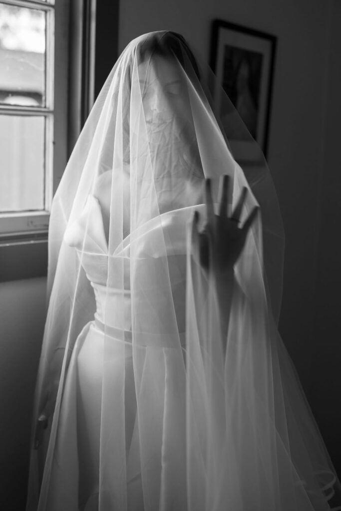 Black and white photo of the bride standing by a window with her veil draped over her face and hand.