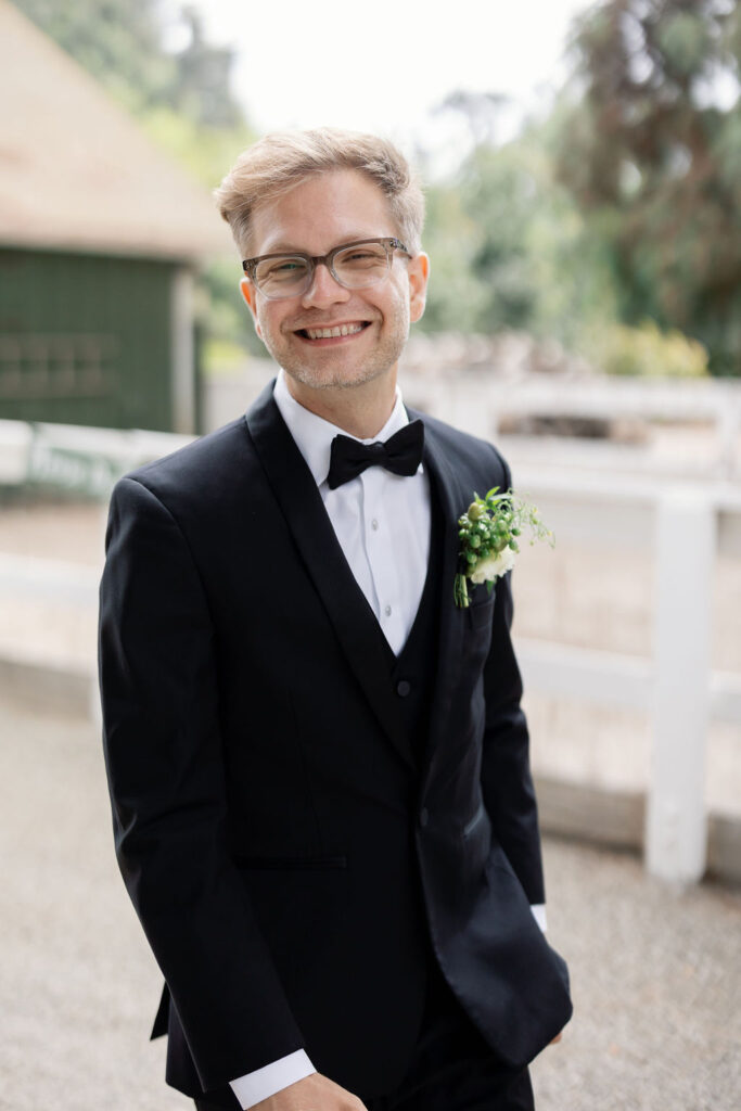 Groom smiling in a black tuxedo with a greenery boutonniere, standing outdoors at McCormick Home Ranch.