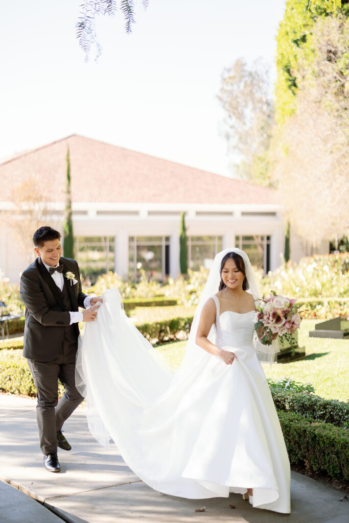 Groom helping bride with her train while walking through the gardens at Nixon Library Yorba Linda wedding.