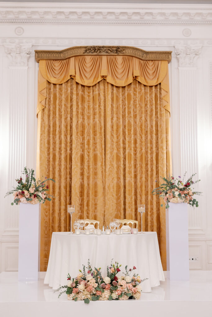 Reception sweetheart table framed by florals and gold drapery in the East Room at the Nixon Library in Yorba Linda.