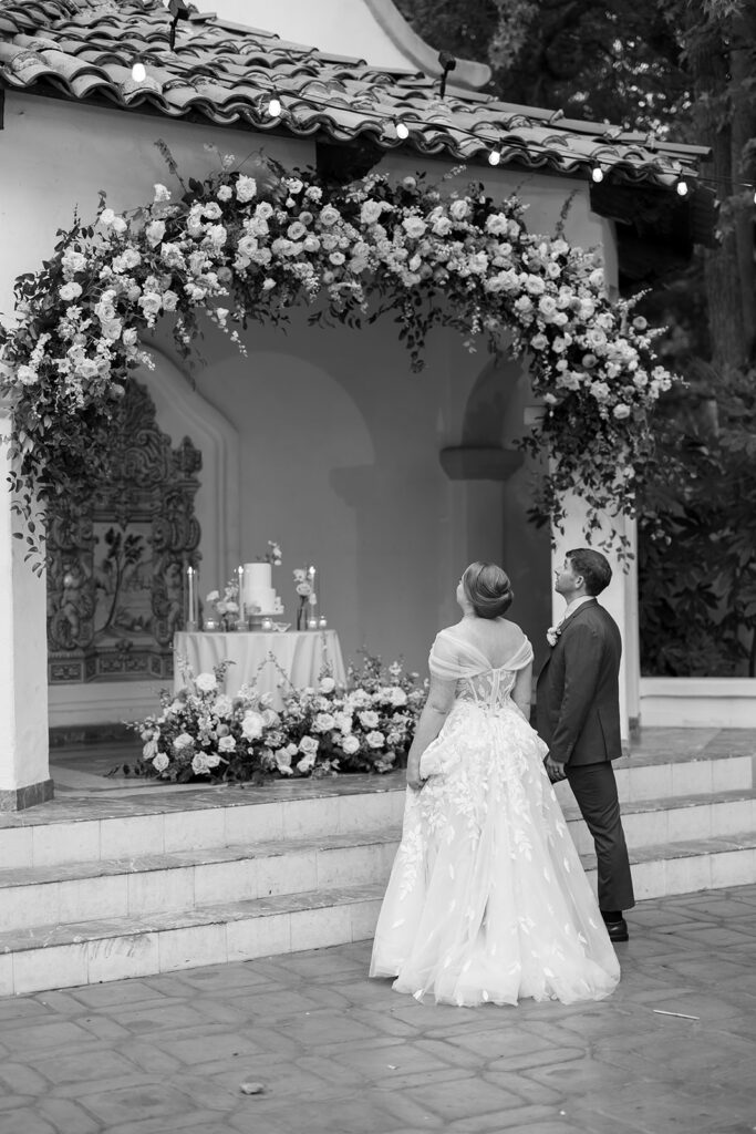 Black and white portrait of the couple standing beneath a floral ceremony arch at El Teatro during their Rancho Las Lomas wedding.