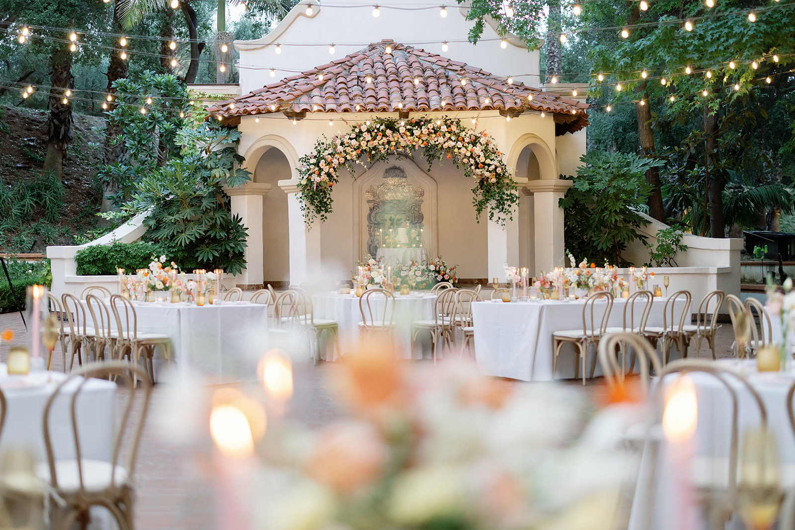 Wide view of El Teatro reception space at Rancho Las Lomas wedding with string lights, round tables, and floral installations.