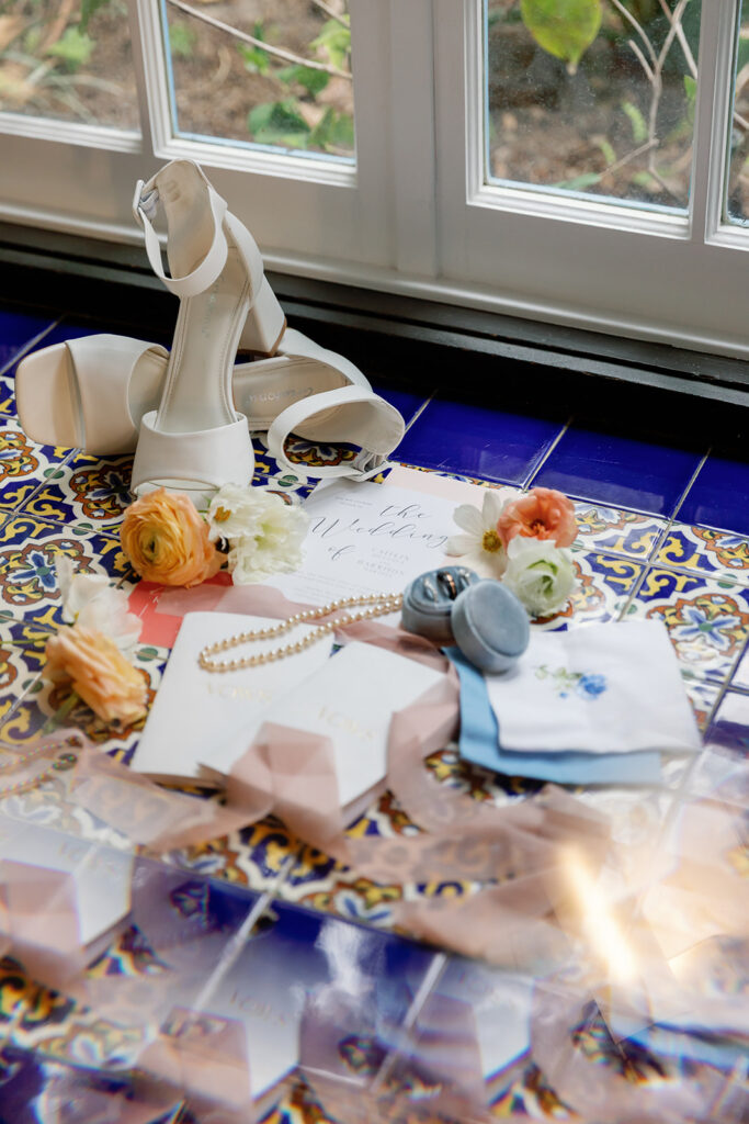 Bride’s white heels, citrus-toned florals, vow books, and invitation suite styled on blue tile during a Rancho Las Lomas wedding.