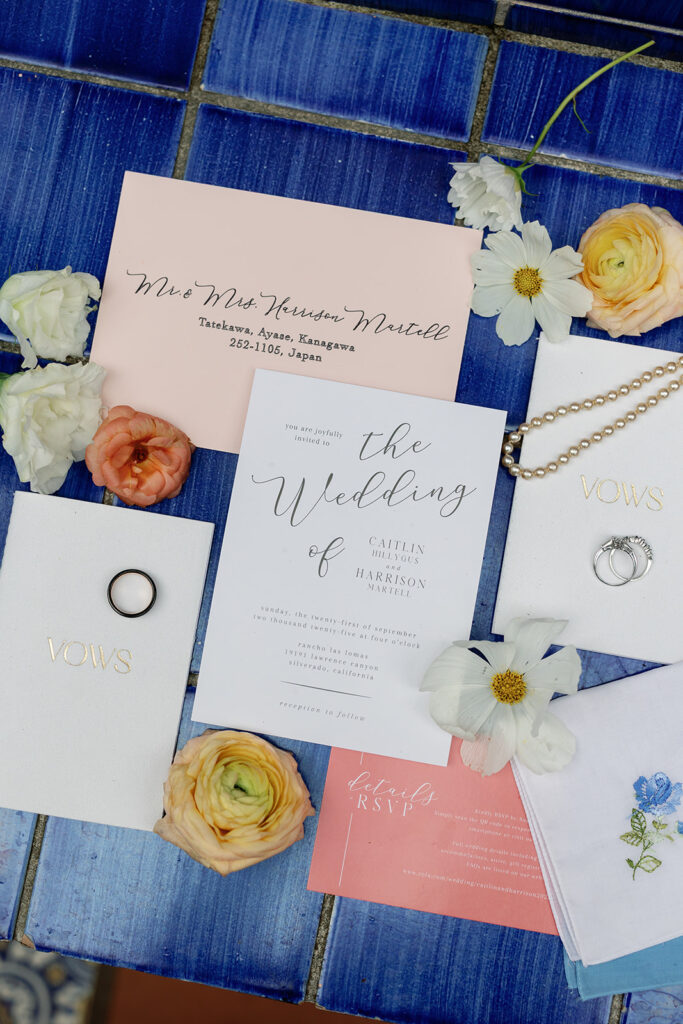 Flat lay of Rancho Las Lomas wedding invitations, rings, and citrus-inspired florals on blue tile.