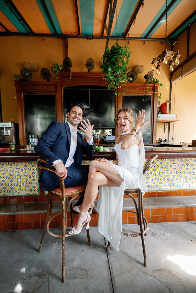 Bride and groom showing wedding rings while sitting at bar inside Habana Irvine Spectrum after courthouse elopement.