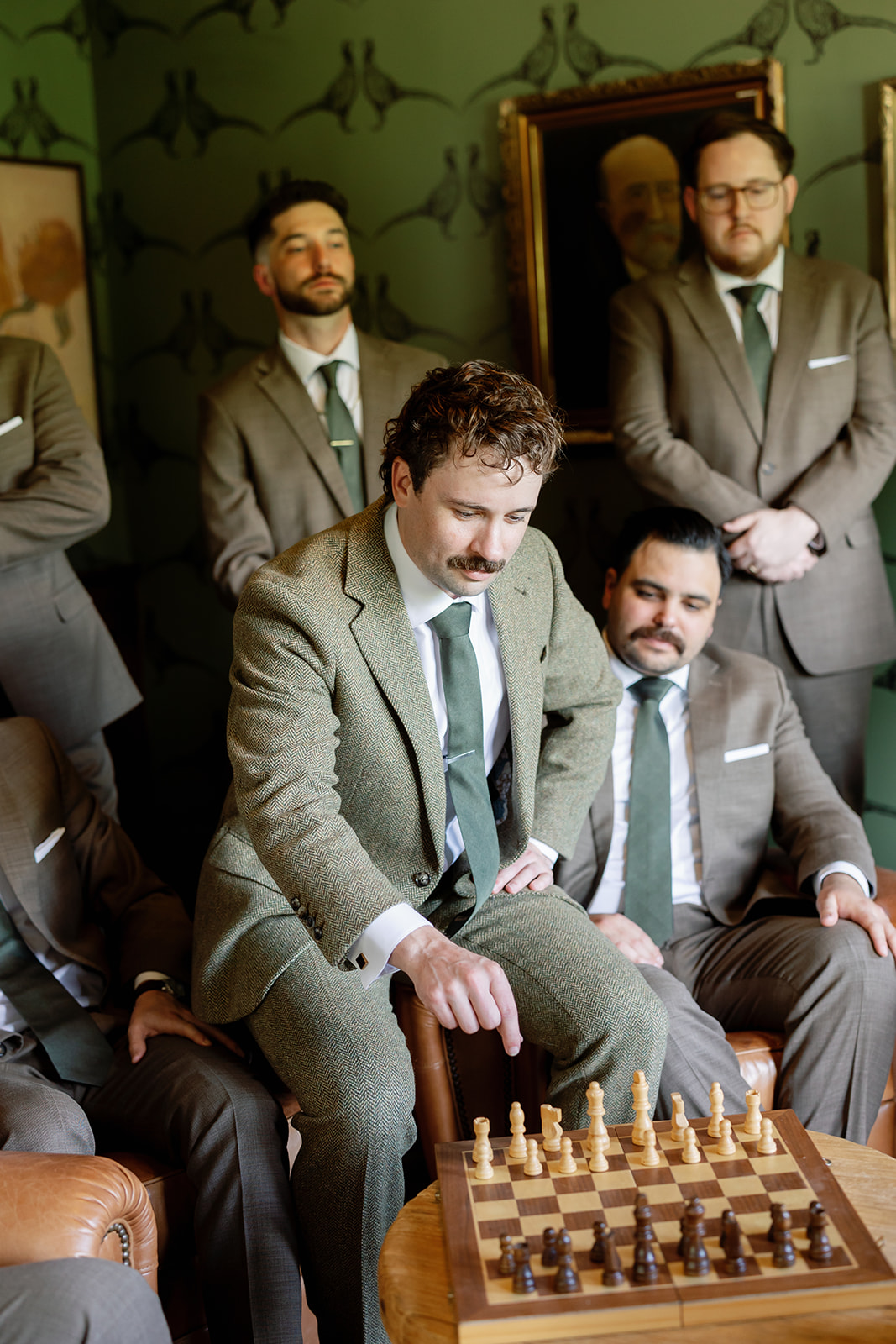 Groom moving a chess piece while surrounded by his groomsmen in green wallpaper room during Swanner House wedding getting ready.