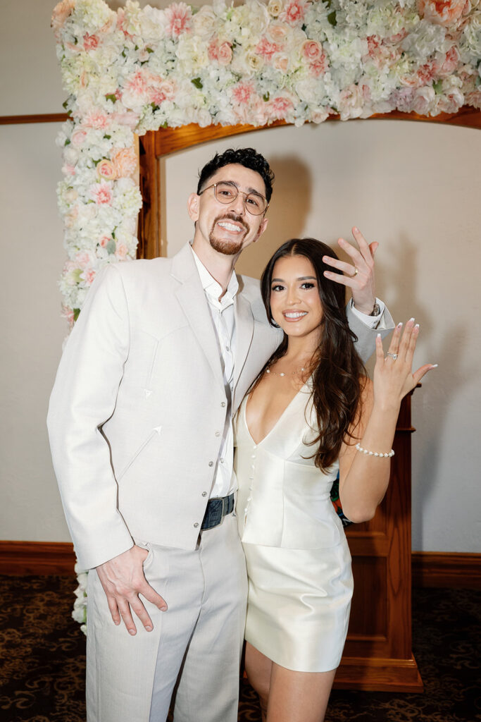 Bride and groom showing wedding rings under floral arch at Old Orange County Courthouse wedding.
