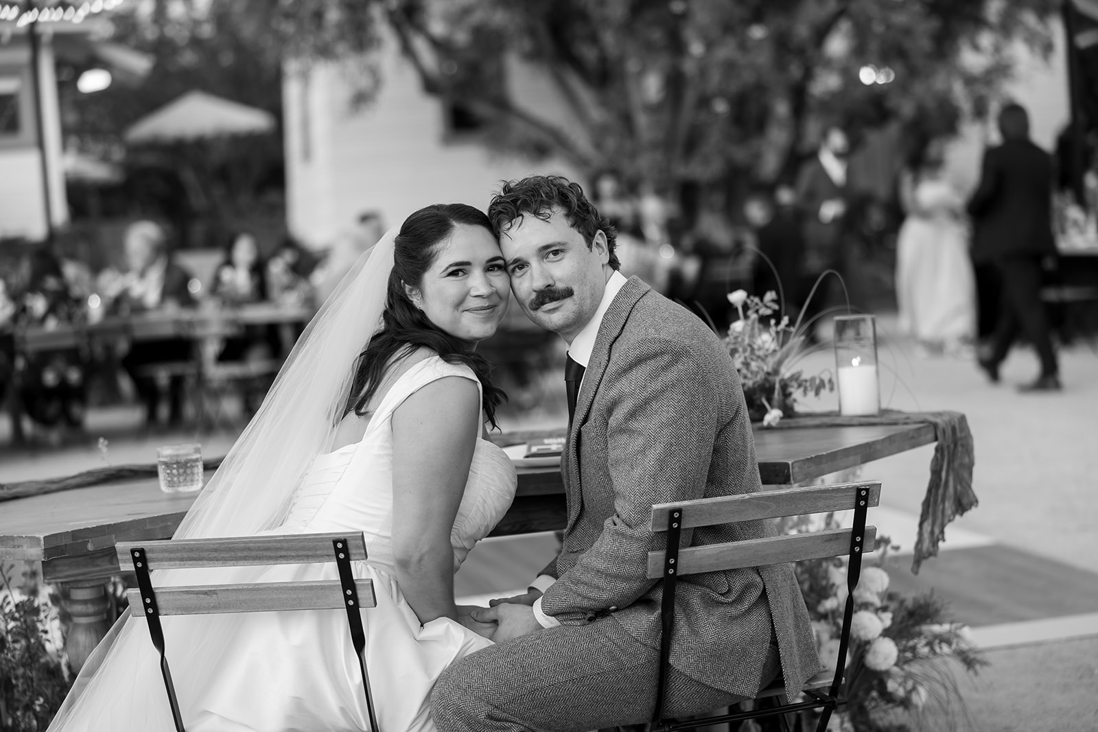 Bride and groom seated together at reception table with guests in the background at a Swanner House wedding in San Juan Capistrano.