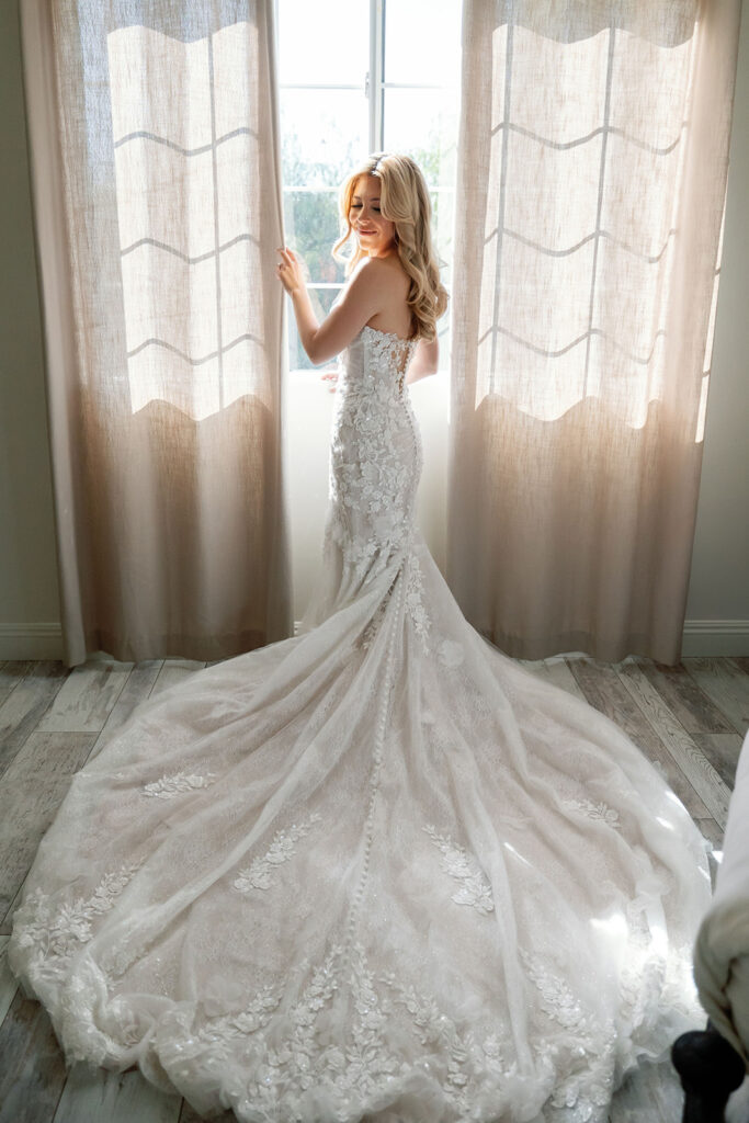 Bride wearing a lace wedding dress in front of the window inside the Lake Oak Meadows getting ready house.