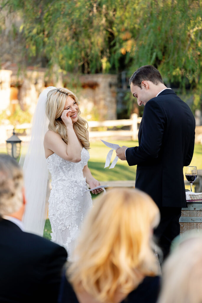 Bride wiping tears while groom reads vows during their Lake Oak Meadows wedding ceremony.