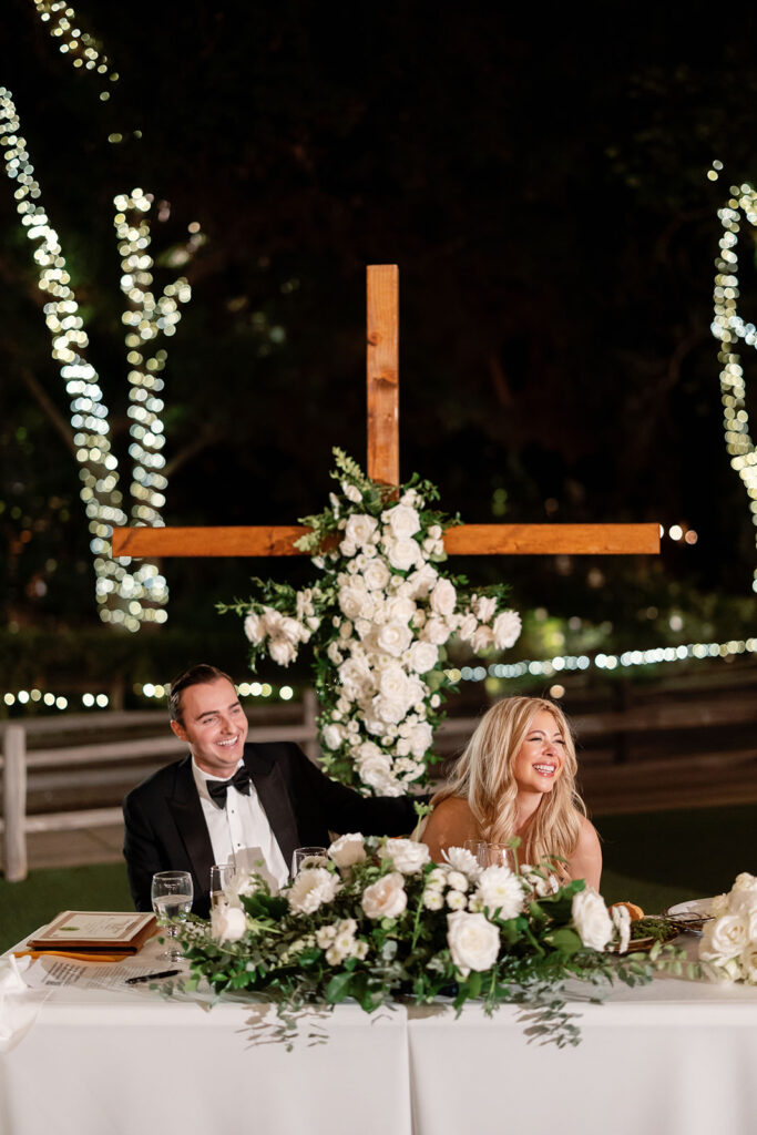 Bride and groom sitting at their sweetheart table with white florals and a floral cross behind them.