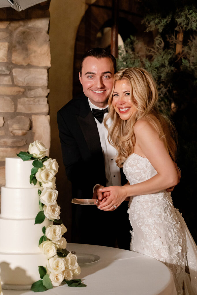 Bride and groom smiling together beside their white wedding cake with rose details.