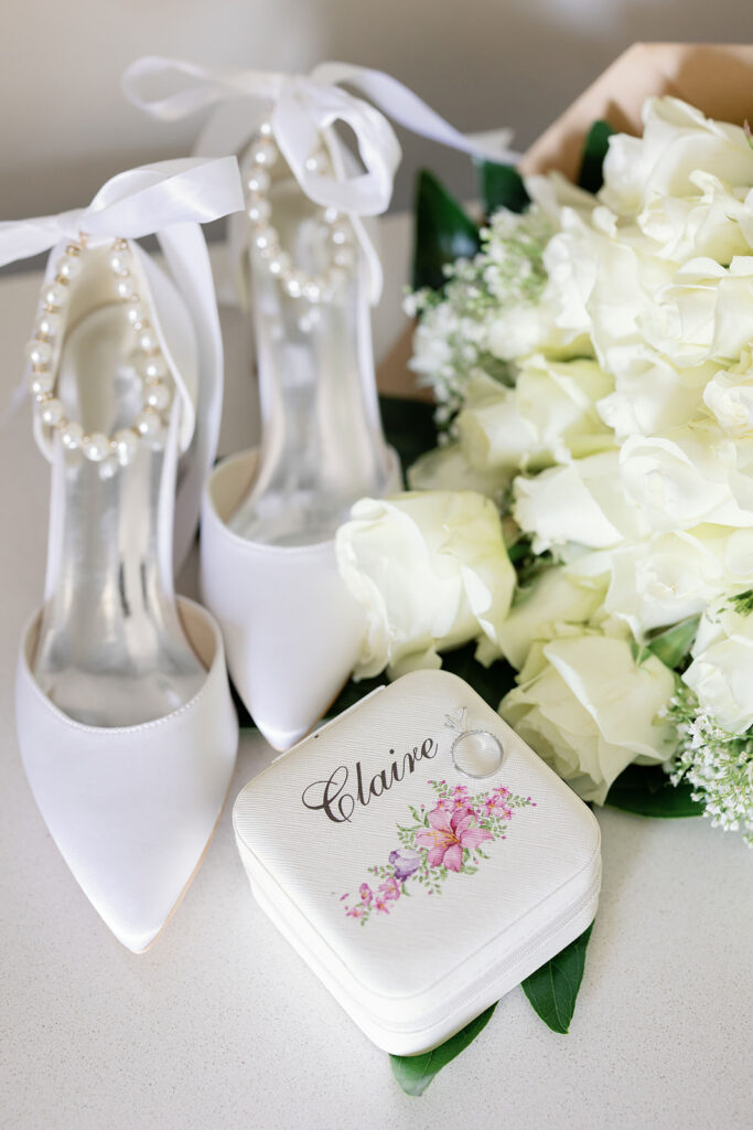 Bridal shoes, wedding rings, white flowers, and a personalized ring box arranged together.
