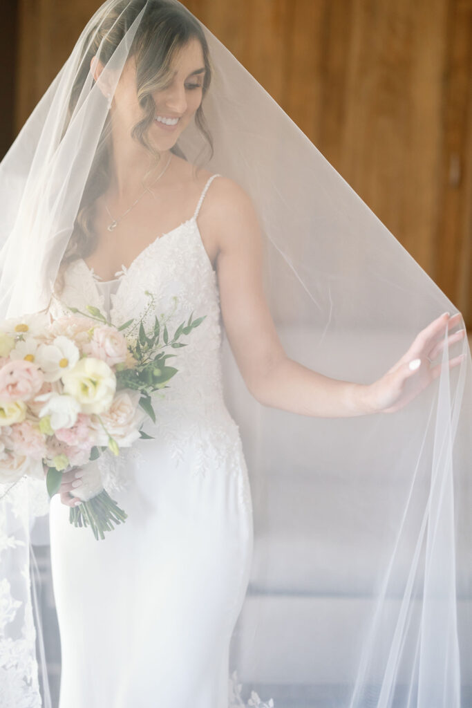 Bride smiling beneath her veil while holding a blush and white wedding bouquet.