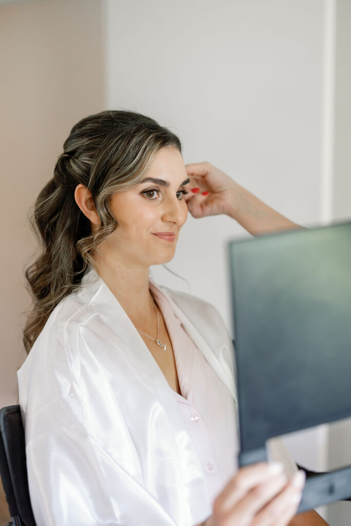 Bride looking in a handheld mirror as she gets her hair and makeup done for her wedding day. 