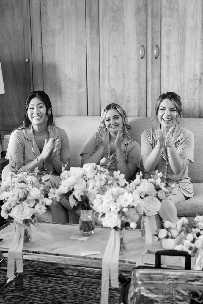 Black and white photo of bridesmaids sitting on a couch and clapping as the bride gets her hair and makeup done.