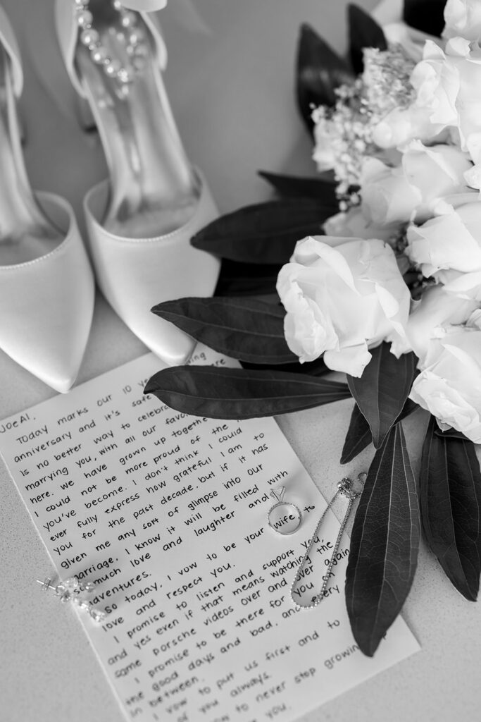 Black and white photo of handwritten vows, rings, florals, and heels. 