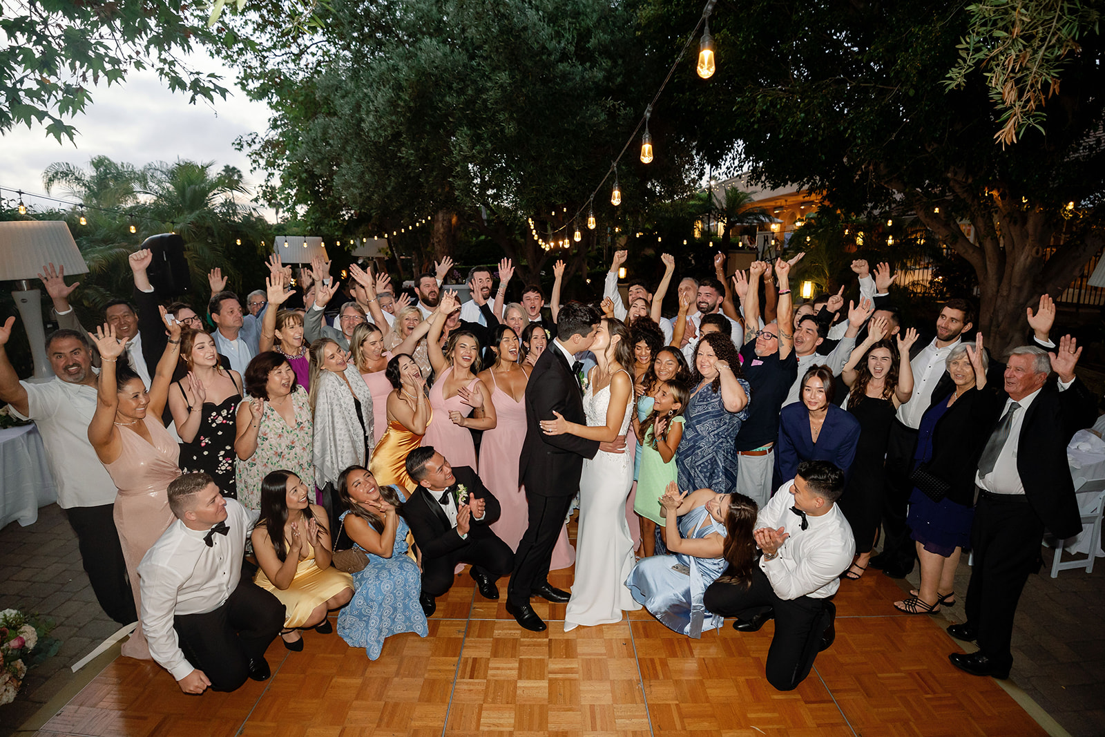 Couple kissing while surrounded by cheering wedding guests on the outdoor dance floor.