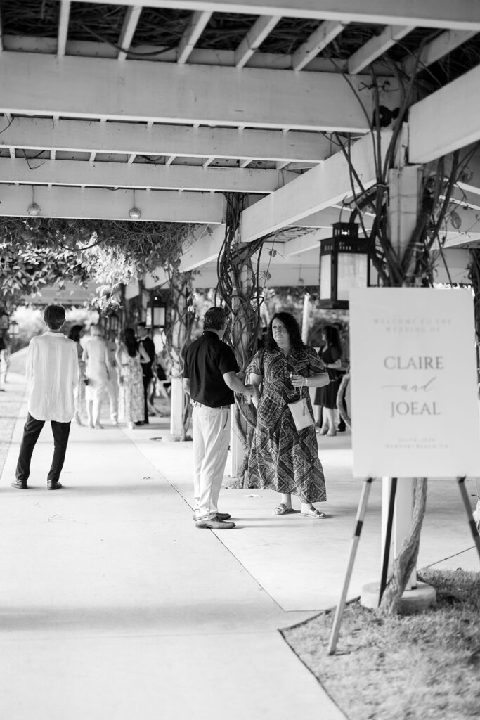 Guests mingling beneath a vine-covered pergola beside a wedding welcome sign.