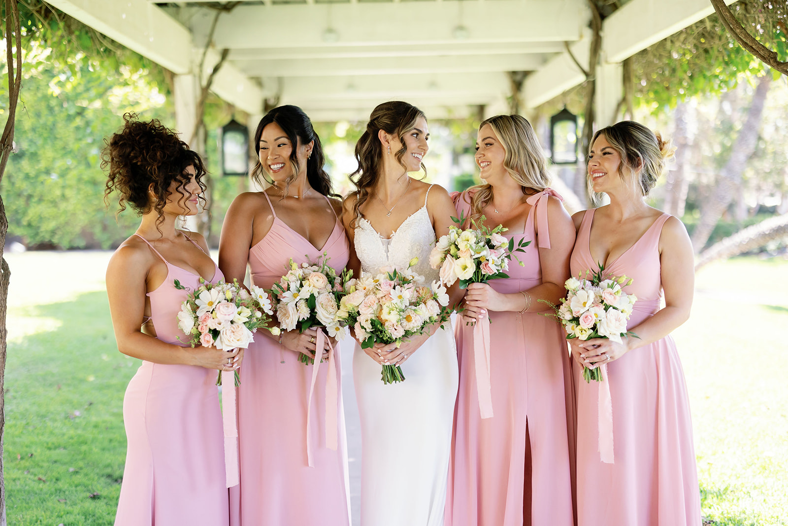 Outdoor photo of a bride and bridesmaids in pink dresses posing under an ivy-covered pergola. 