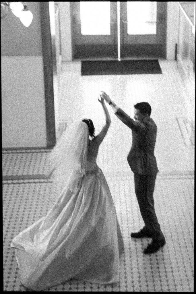 Black and white film photo of a groom twirling his bride inside the Old Orange County Courthouse in Santa Ana, California.
